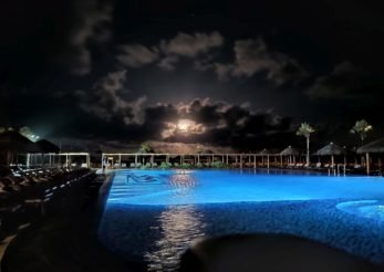 #PraCegoVer Audiodescrição: fotografia noturna com uma piscina iluminada de azul em primeiro plano, uma lua semi encoberta por nuvens